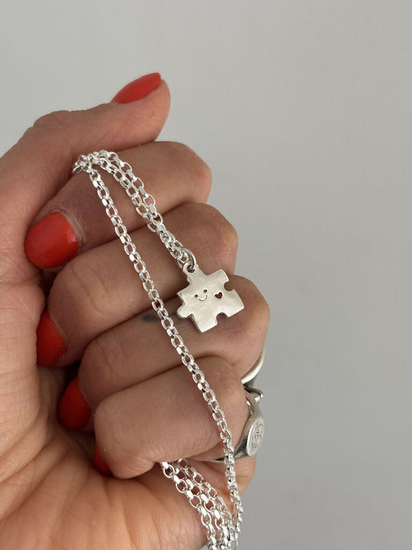 Hand holding a puzzle charm necklace the charm is a puzzle with a smiley face and red heart