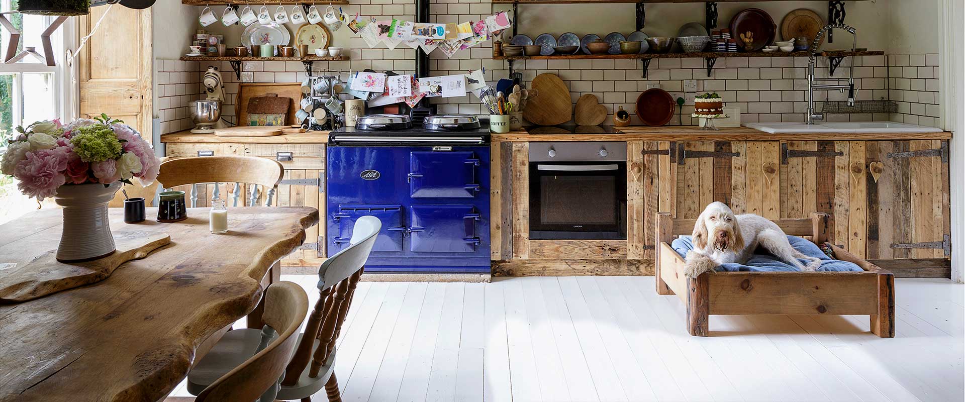 A rustic kitchen interior with wooden cabinets, a blue Aga stove, white subway tiles, and a white dog lying on a wooden pet bed. A large wooden dining table with flowers is visible in the foreground.