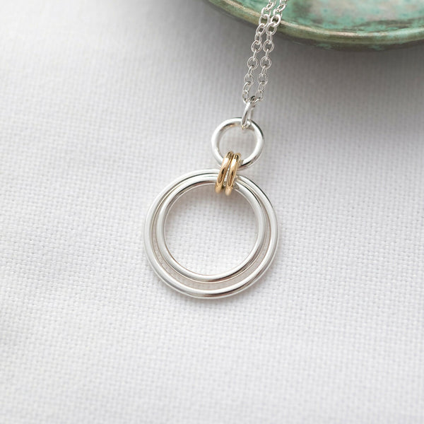 A gold and silver two ring circle necklace displayed on white material with a green jewellery dish in the background.