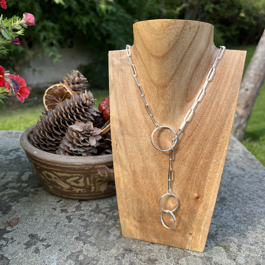 Handmade sterling silver lariat necklace on a chunky silver paperclip chain, displayed on a wooden bust with a natural background