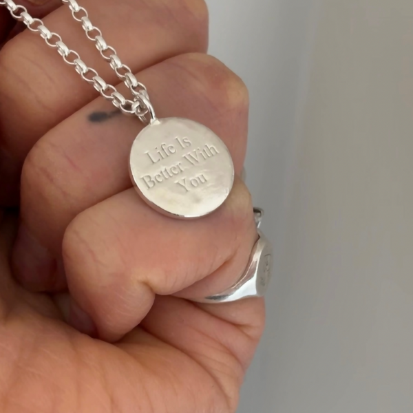 Hand holding a charm necklace that is engraved with “life is better with you”