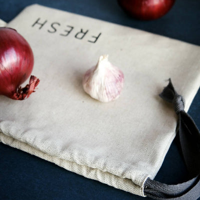 A linen produce bag printed with the word fresh, shown with garlic and onions.