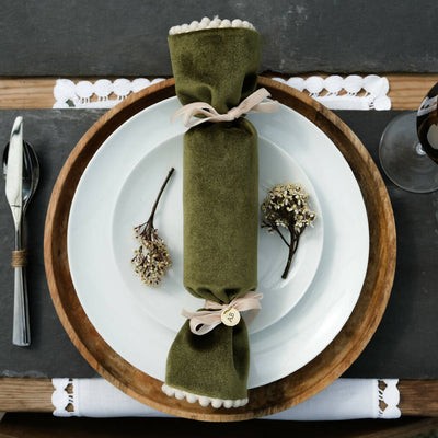 A green velvet christmas cracker with pom pom trim set out on a dinner plate.